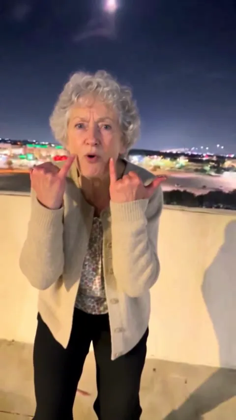 AI video: a white-haired grandmother in a beige cardigan throws rock horns and sings the emo Choosin' Texas on a parking garage roof at night