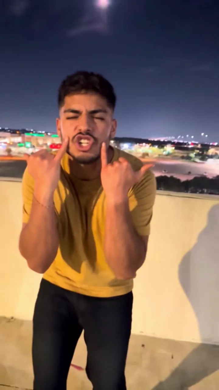 AI video: a young man in a mustard T-shirt throws rock horns and sings the emo Choosin' Texas on a parking garage roof at night