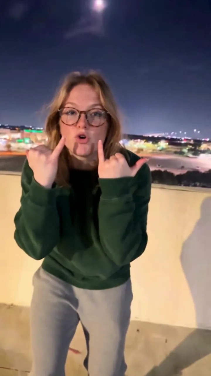 AI video: a young woman with glasses and a green sweater throws rock horns and sings the emo Choosin' Texas on a parking garage roof at night